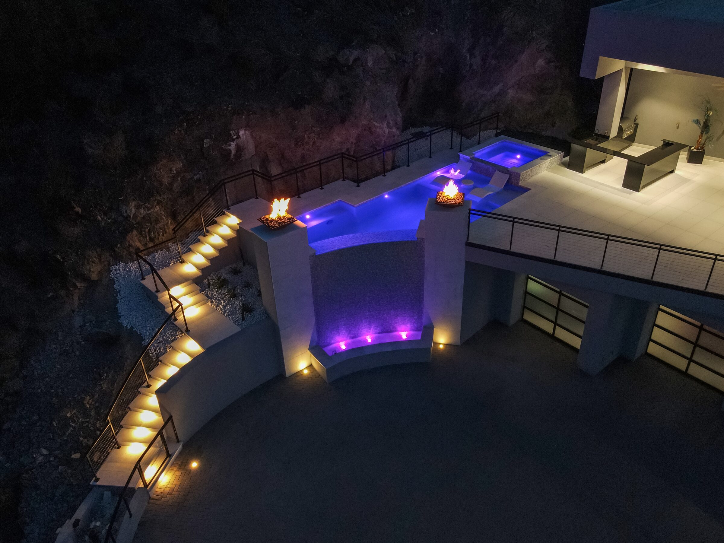 Dramatic nighttime view of Paradise Valley hillside pool with 16-foot negative edge wall, fire bowls, marine-grade steel railing, and custom illuminated staircase